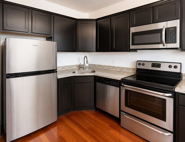 A kitchen with dark cabinets, stainless steel appliances, a granite countertop, and a wooden floor.