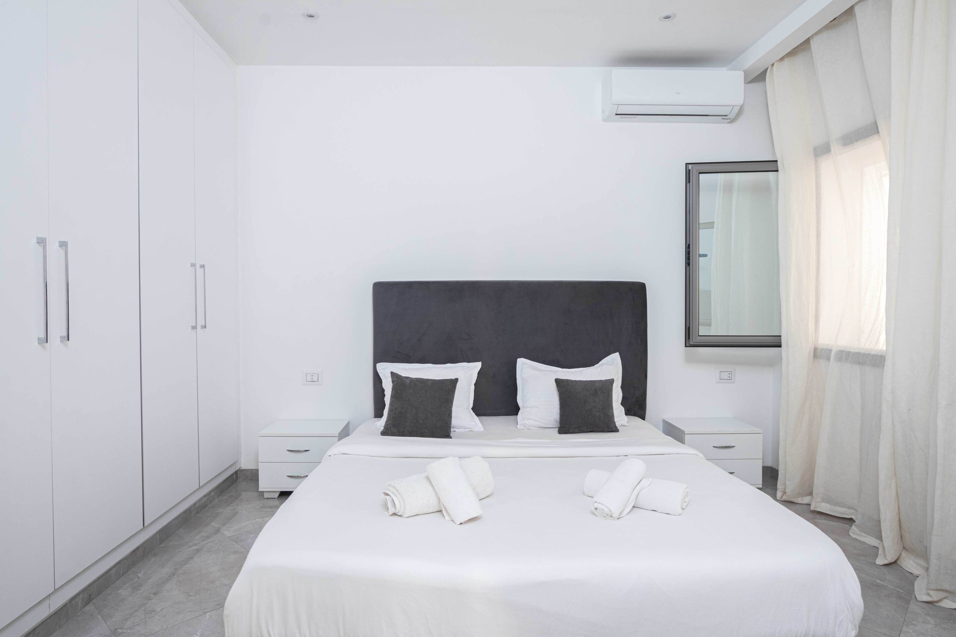 A modern hotel room with a large bed, white walls, a dark headboard, and a window with sheer curtains.