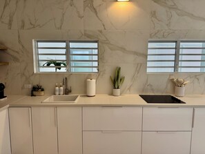 Eine moderne Küche mit Marmorfliesenspiegel, zwei Fenstern mit Jalousien, einem Spülbecken und einer Arbeitsplatte mit Pflanzen und Küchenutensilien.