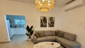 Un salon moderne avec un canapé d’angle, une table basse ronde, une plante en pot et une cuisine en arrière-plan.