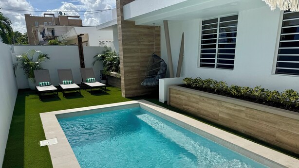 Une piscine dont le bord est recouvert de carreaux, entourée de gazon synthétique et agrémentée d’un bac à fleurs en bois où sont plantées de petites plantes.