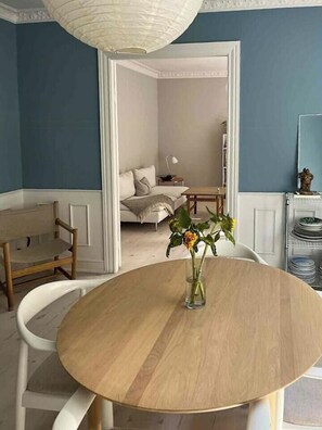 Un comedor con una mesa redonda, sillas, un florero con flores y vista a otra habitación con una cama y una lámpara.
