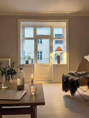 Ein Zimmer mit einem großen Fenster, einem Tisch mit Kerze, einer Lampe und einem Stuhl.