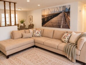 A living room with a beige sectional sofa, a patterned rug, a dining area with a table and chairs, and a large cityscape painting on the wall. - Port Richey Palms (Port Richey)