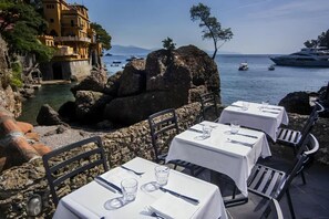 Outdoor dining tables with white tablecloths, set for two, overlooking a rocky coastline with a building and boats in the background.