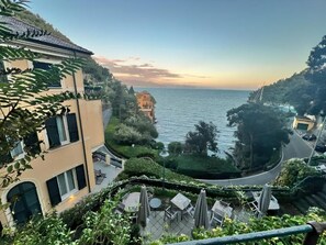 A coastal hotel with a terrace overlooking the sea, a winding road, and lush greenery.