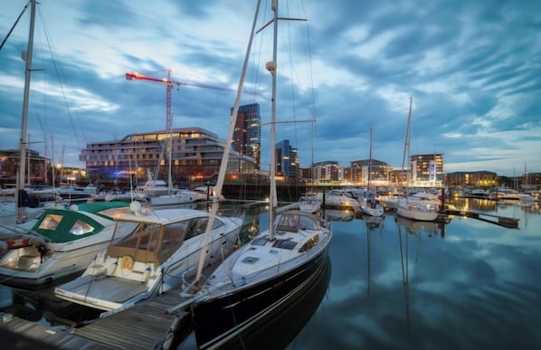 A marina with multiple boats docked, a building with a crane, and a city skyline in the background.