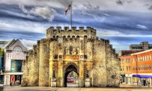 A historic stone castle gate with a flag, surrounded by modern buildings.