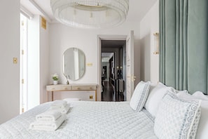 Ein Hotelzimmer mit Bett, Schminktisch, Spiegel, einer Chandelüster und Blick auf den Flur.