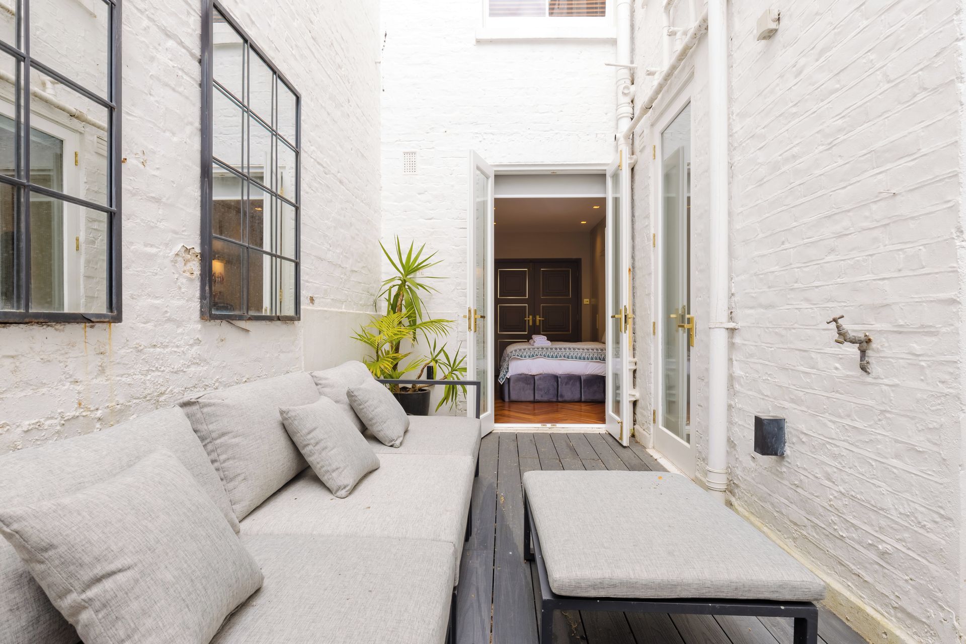 A narrow outdoor space with a white brick wall, a wooden bench, a cushioned seating area, and a door leading to a bedroom.