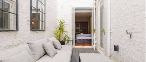 A narrow outdoor space with a white brick wall, a wooden bench, a cushioned seating area, and a door leading to a bedroom.