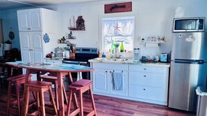 Eine Küche mit Holztisch, Barhocker, Kühlschrank und einem Fenster mit einer Meerfarn-Dekoration.