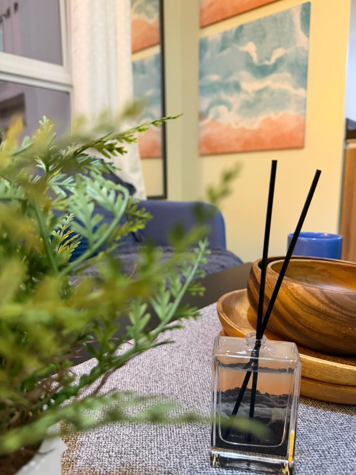A small glass container with black straws, a wooden bowl, and a plant on a textured surface.