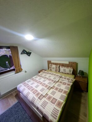 A bedroom with a bed, a nightstand, a window with curtains, a radiator, and a wall-mounted clock.
