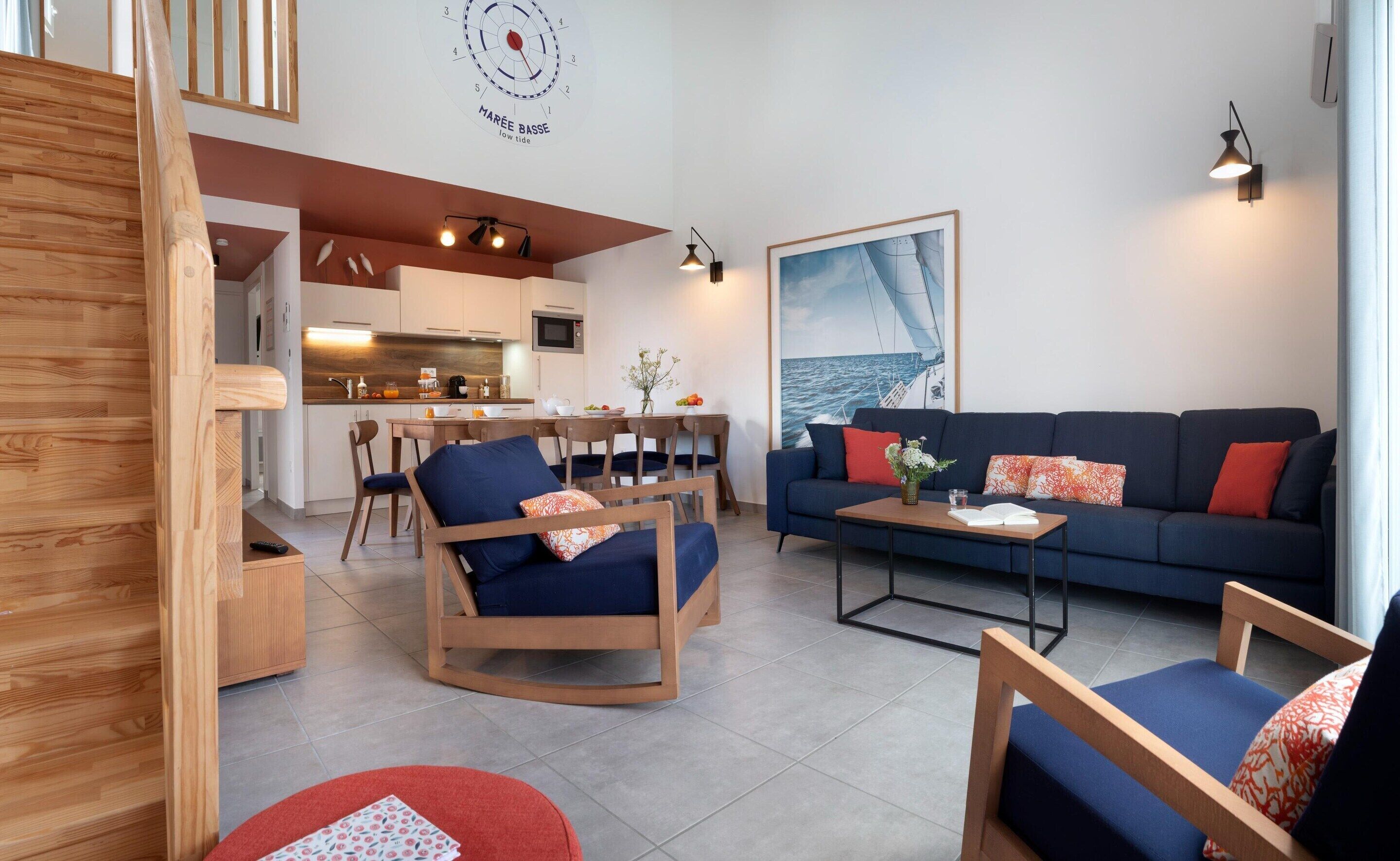 Ein modernes Wohnzimmer mit einer Couch, Sesseln, einem Couchtisch und einem Essbereich, im Hintergrund ist eine Küche zu sehen.