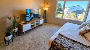 A bedroom with a bed, a TV on a stand, a window with a view of buildings, and two potted plants. - Refugio Urbano  Bariloche (San Carlos de Bariloche)