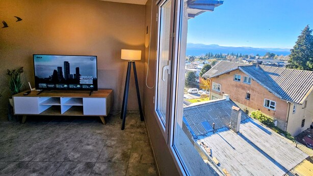 A room with a TV on a stand, a floor lamp, and a window overlooking a residential area. - Refugio Urbano  Bariloche (San Carlos de Bariloche)