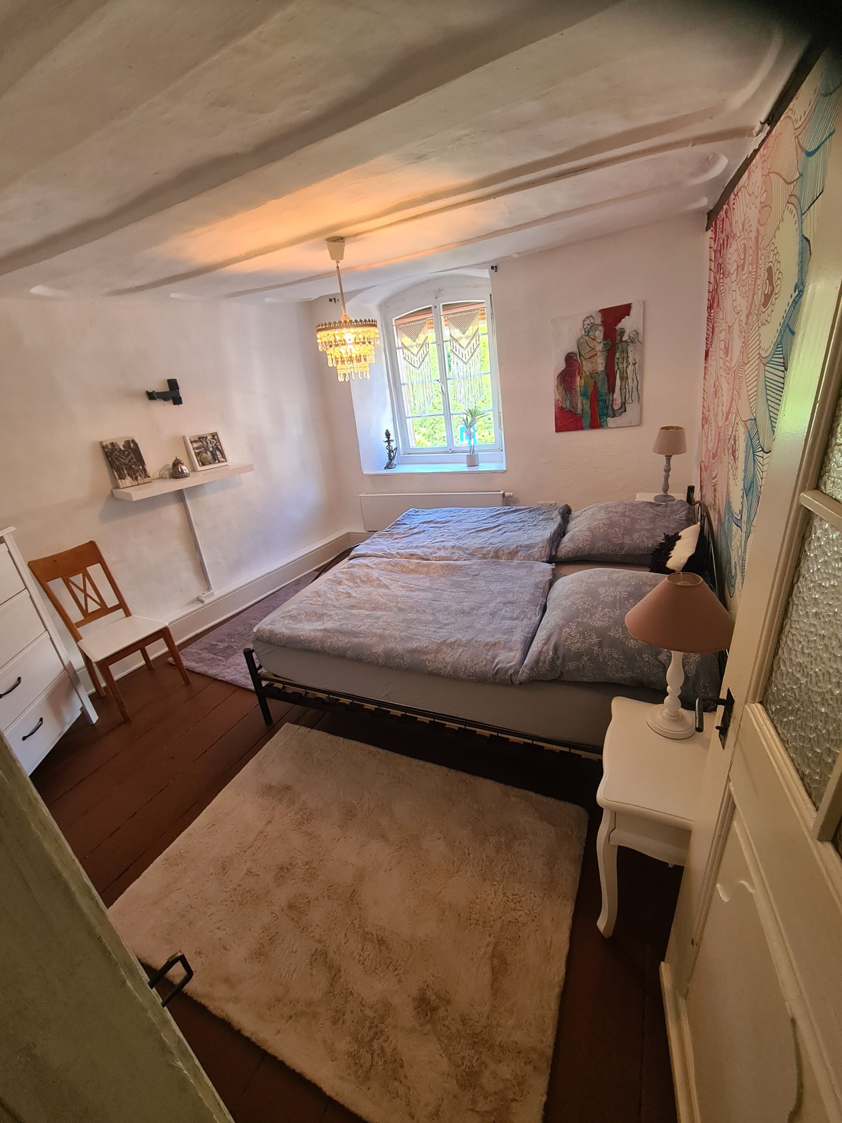 Um quarto com cama, janela, lustre, abajur e um quadro.