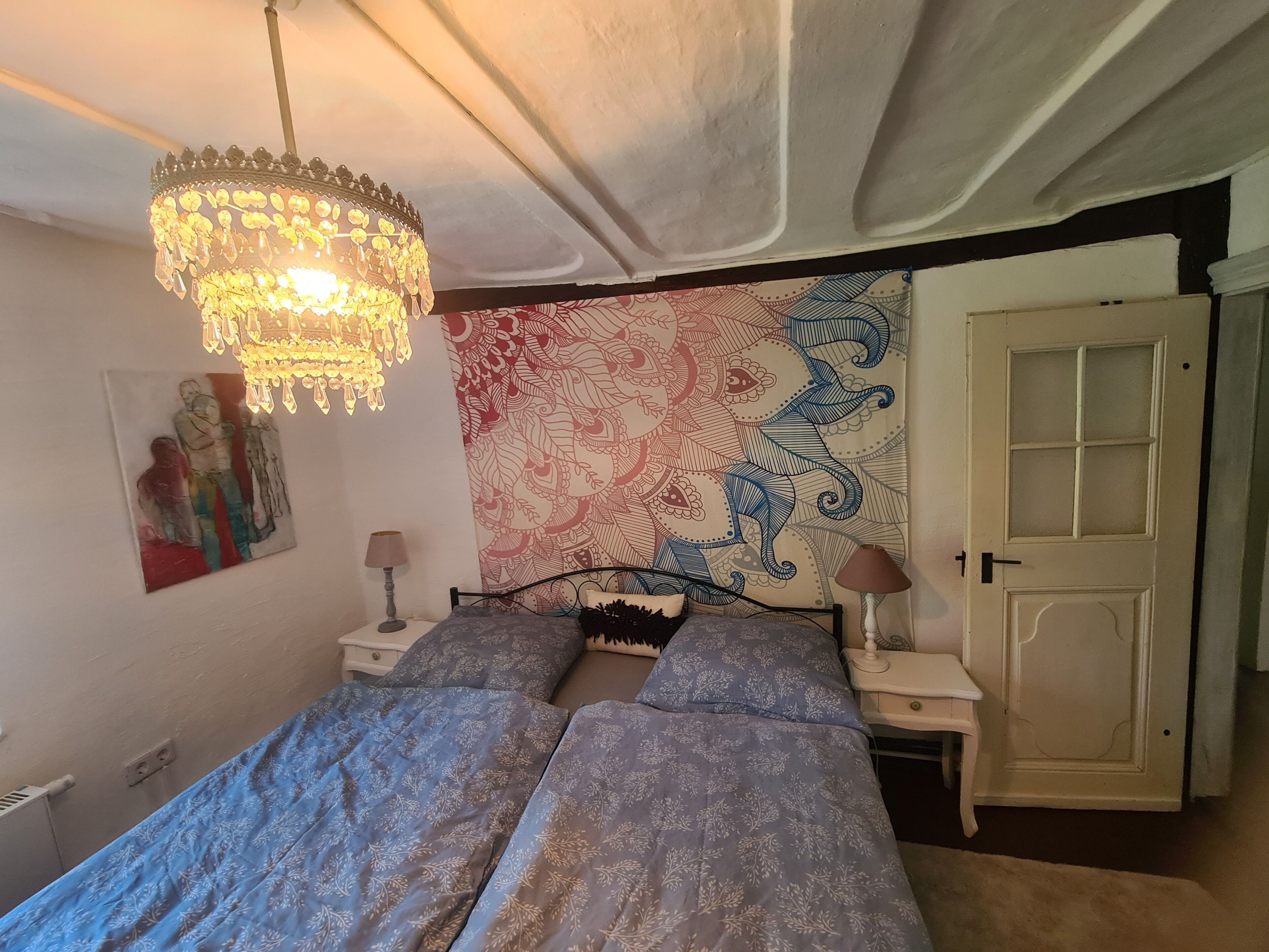 Un dormitorio con una cama, dos mesitas de noche, una araña, un tapiz y una puerta.