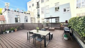 Une terrasse en bois avec une table et des chaises, entourée de plantes en pot et d’un barbecue.