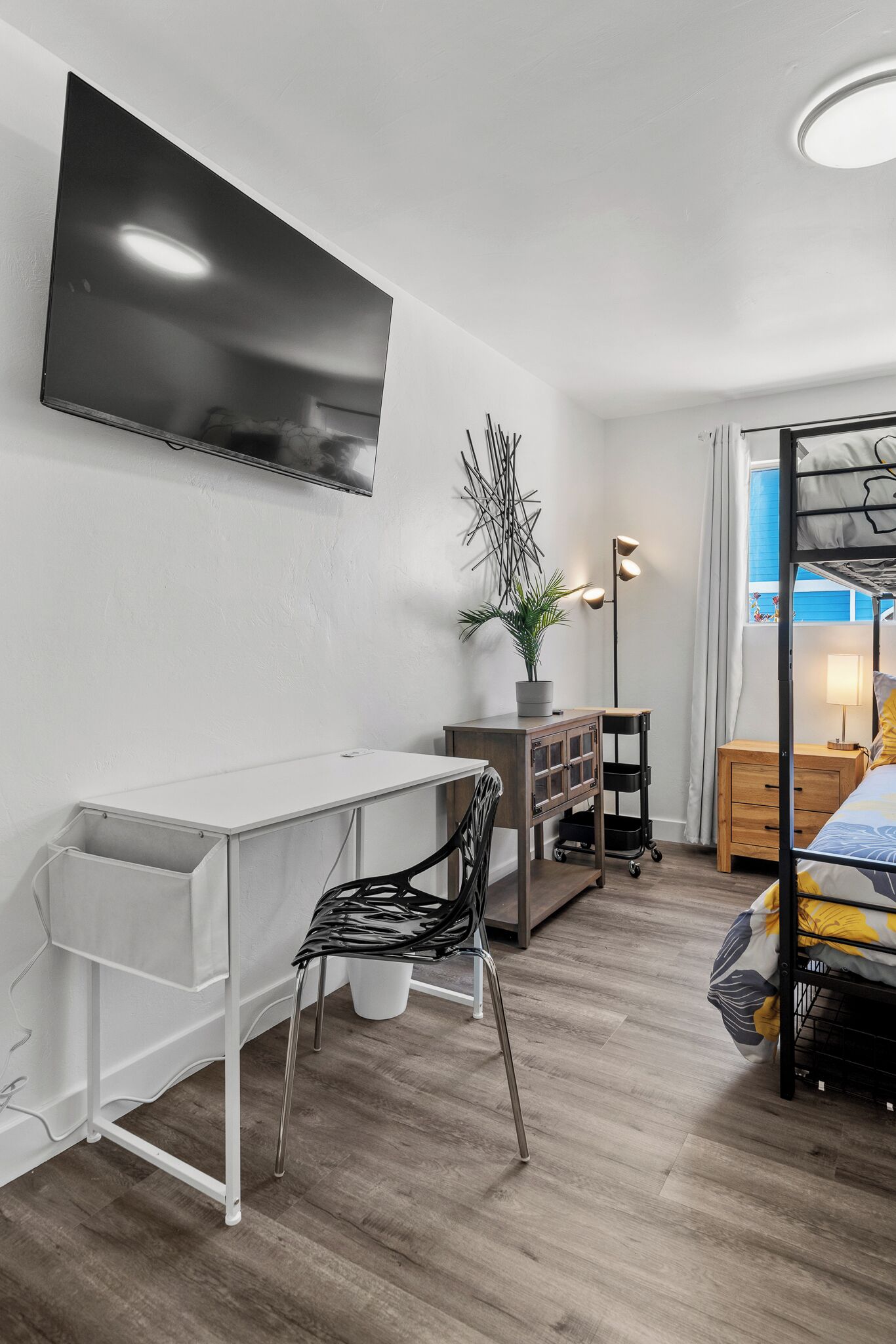 Une chambre moderne équipée d’un lit, d’un bureau, d’une chaise, d’un téléviseur, d’une plante en pot, d’une lampe et d’une fenêtre avec des rideaux.