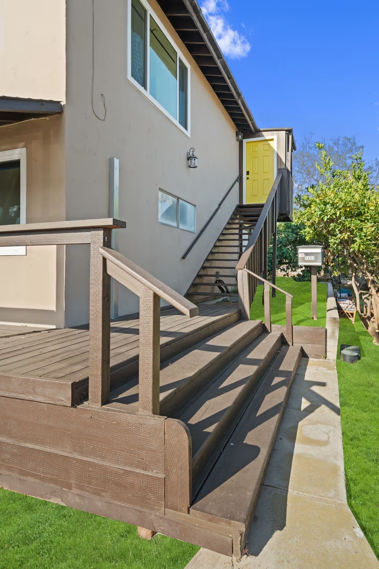 A building with a staircase leading to a yellow door, a wooden deck, and a well-maintained lawn.