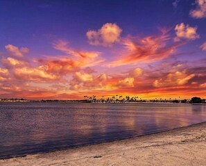 Ein Strand mit einem Sonnenuntergangshimmel, ruhigem Wasser und einer fernen Küstenlinie mit Palmen.