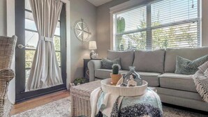 Un salon comprenant un canapé, un fauteuil en osier, une fenêtre avec des stores, une lampe et un plateau décoratif sur lequel sont disposés divers objets.