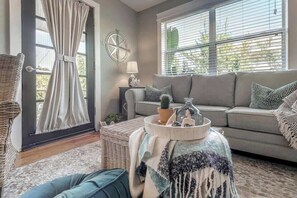 Ein Wohnzimmer mit einer Couch, einem Korbsessel, einem Fenster mit Jalousien, einer Lampe und einem dekorativen Tablett mit Gegenständen.