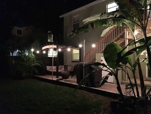 Ein Nachtblick auf ein Haus mit überdachter Terrasse, Gartenmöbeln und einer Treppe, die zu den oberen Stockwerken führt.