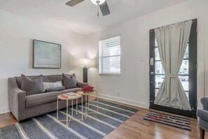 Ein Wohnzimmer mit einer Couch, einem kleinen Tisch, einer Lampe, einem Fenster mit Jalousien und einer Tür mit Vorhängen.