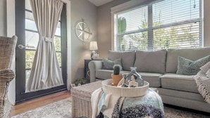 Un salon comprenant un canapé, un fauteuil en osier, une fenêtre avec des stores, une lampe et un plateau décoratif sur lequel sont disposés divers objets.