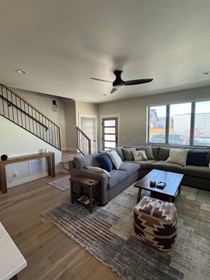A spacious living room with a staircase, a ceiling fan, and a large window with a view of buildings outside. - Cozy mountain townhome in beautiful Salida (Salida)