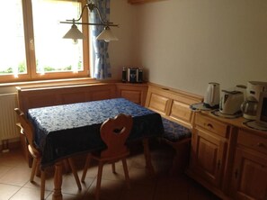 A kitchen with a table and chairs, wooden cabinets, a window with curtains, and kitchen appliances. - Chalet Bergblick in Sachrang Bavaria for 4 Guests - Experience Pure Nature! (Aschau im Chiemgau)
