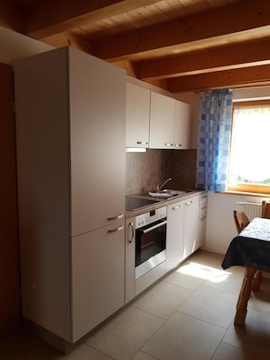 A compact kitchen with a sink, stove, and cabinets, a dining table with a tablecloth, and a window with curtains. - Chalet Bergblick in Sachrang Bavaria for 4 Guests - Experience Pure Nature! (Aschau im Chiemgau)