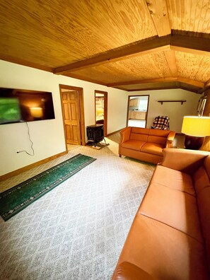 A cozy living room with a wooden ceiling, a television mounted on the wall, a sofa, a chair, and a lamp.