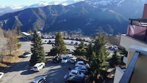 A parking lot with cars, a bus, and a building with a balcony overlooking a mountainous landscape. - Panoramic view and mountain cocoon Pra-Loup 1500 (Uvernet-Fours)