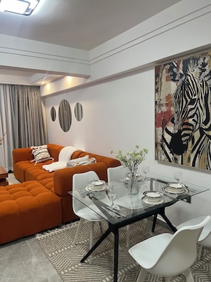 Ein modernes Wohnzimmer mit einem Esstisch aus Glas, gedeckt für zwei Personen, einer Couch und einem großen, abstrakten Gemälde an der Wand.
