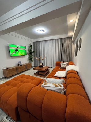 Ein modernes Wohnzimmer mit einem großen, orangefarbenen Ecksofa, einem Holz-TV-Schrank, einem Flachbildfernseher und einem großen Fenster mit Vorhängen.