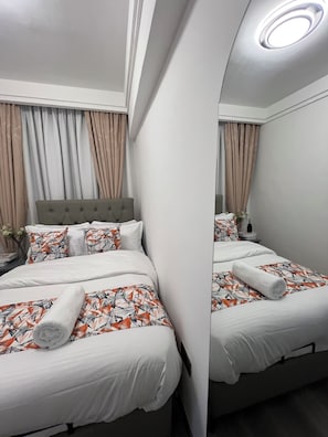 Ein Hotelzimmer mit zwei Betten, einem grauen Kopfteil, weißer Bettwäsche und einem Spiegel, der den Raum reflektiert.