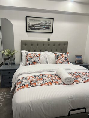 En pænt redt seng med mønstret sengetøj, et sengegærde, et natbord med en vase blomster og et indrammet billede på væggen.