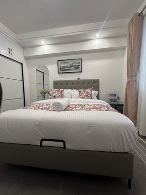 En pænt redt seng med sengegavl, et natbord med lampe, et billede i ramme på væggen og et vindue med gardiner.