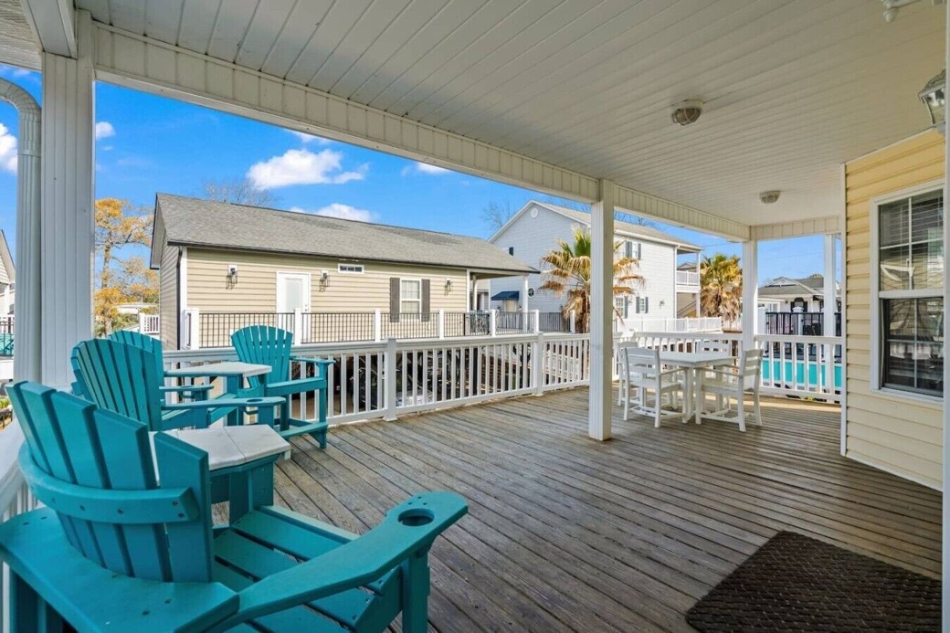 A covered outdoor deck with wooden flooring, white railings, and blue Adirondack chairs.