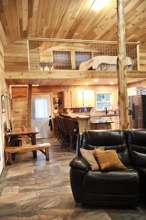 Interior de una cabaña de madera con comedor, cocina y un sofá de cuero negro.