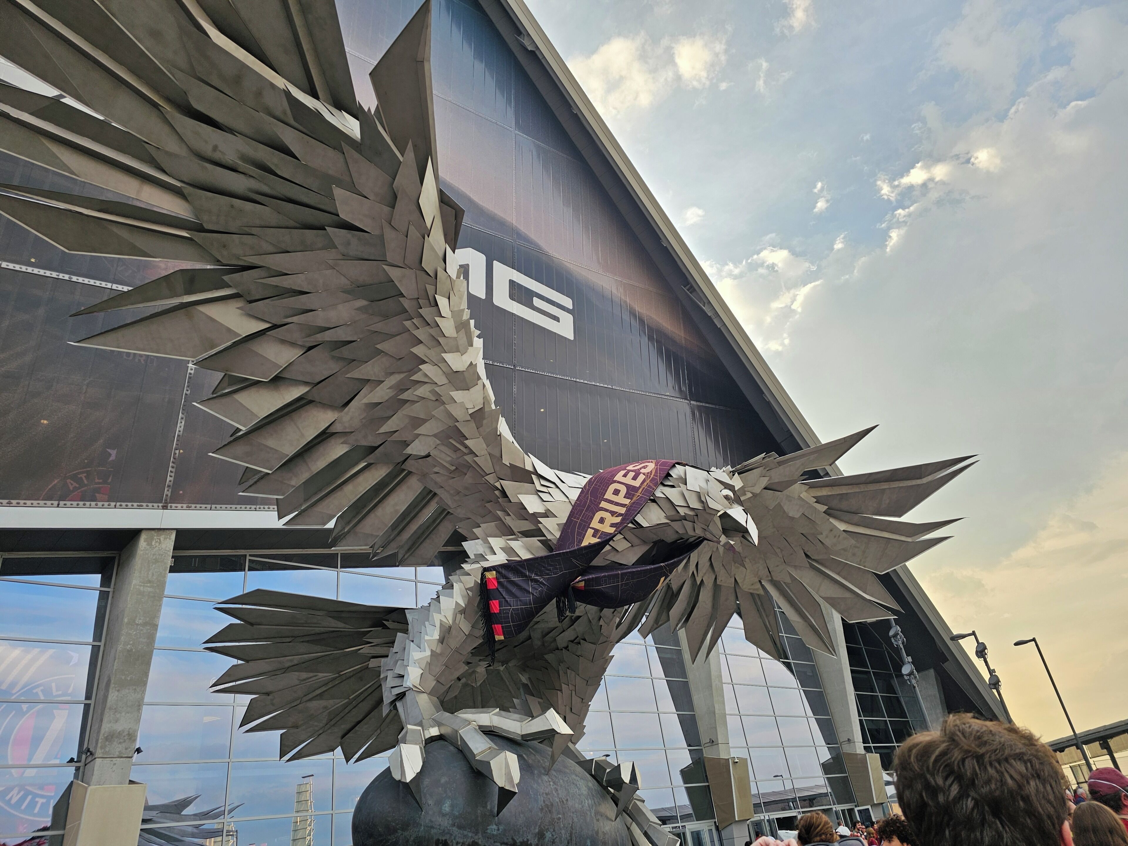 A large metallic sculpture resembling an eagle with outstretched wings, positioned in front of a modern building with a glass facade.