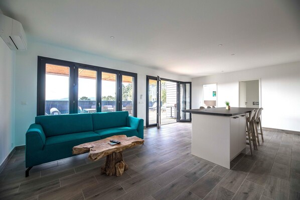 A modern living room with a teal sofa, a wooden coffee table, and a kitchen area with a bar and stools. - Lecci Villa With Outdoor Pools (Lecci)