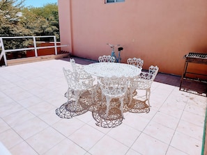 A round outdoor table with white chairs, a blue bicycle, and a grill on a tiled patio.
