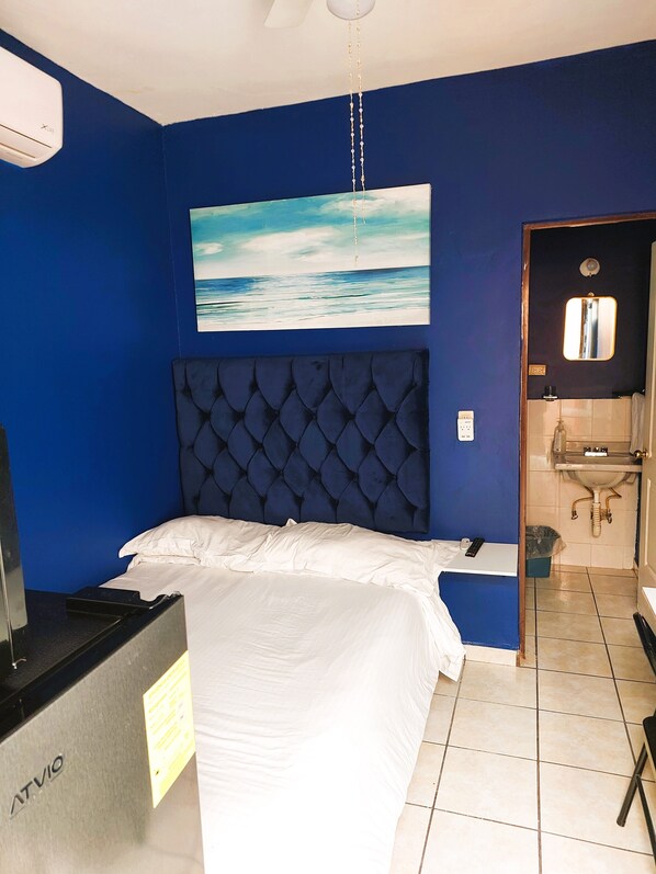 A room with a bed, a painting of a beach, a blue headboard, a TV, and a bathroom visible through an open door.