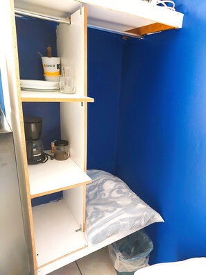 Une petite piĂšce, de taille modeste, avec un mur bleu, un lit dotĂ© dâun matelas blanc, une petite Ă©tagĂšre sur laquelle se trouvent une tasse et un pot, et une poubelle.