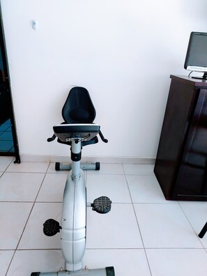 A stationary exercise bike in a room with a chair, a desk with a computer, and a tiled floor.